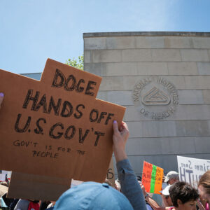 Image of a protest in front of a grey stone building bearing the logo of the NIH. The nearest protester is holding a sign that reads in part, "DOGE hands off US gov't".