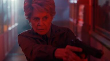 older woman with short gray hair looking grim while pointing a revolver